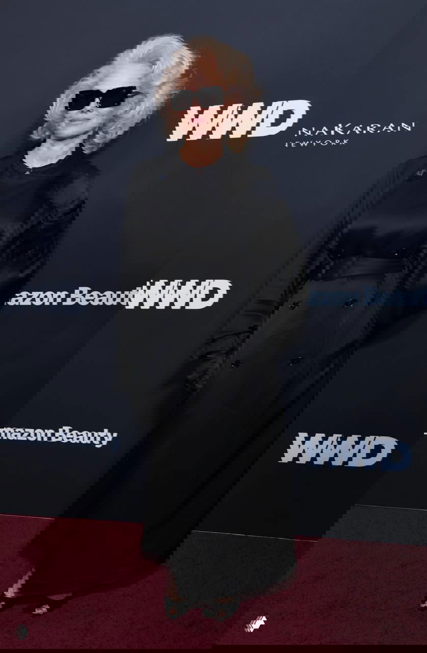 Памела Андерсон с платиновым блондом в чёрном пальто/платье и солнцезащитных очках на красной дорожке WWD Style Awards, общий план на фоне бренд-стены.