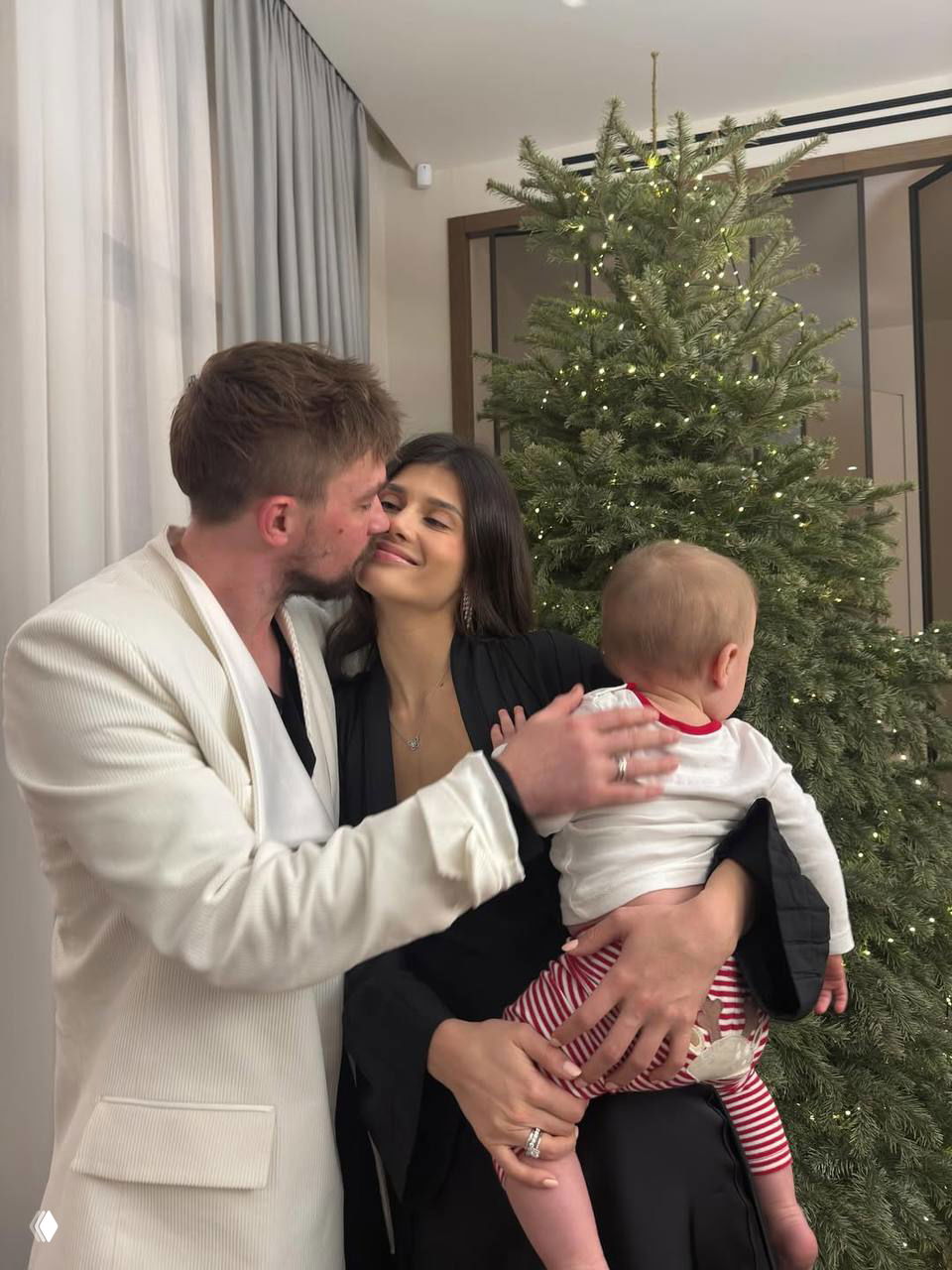 Александр целует Викторию, она держит малыша у украшенной огоньками рождественской ёлки в тёплой домашней атмосфере праздника и семейного уюта.