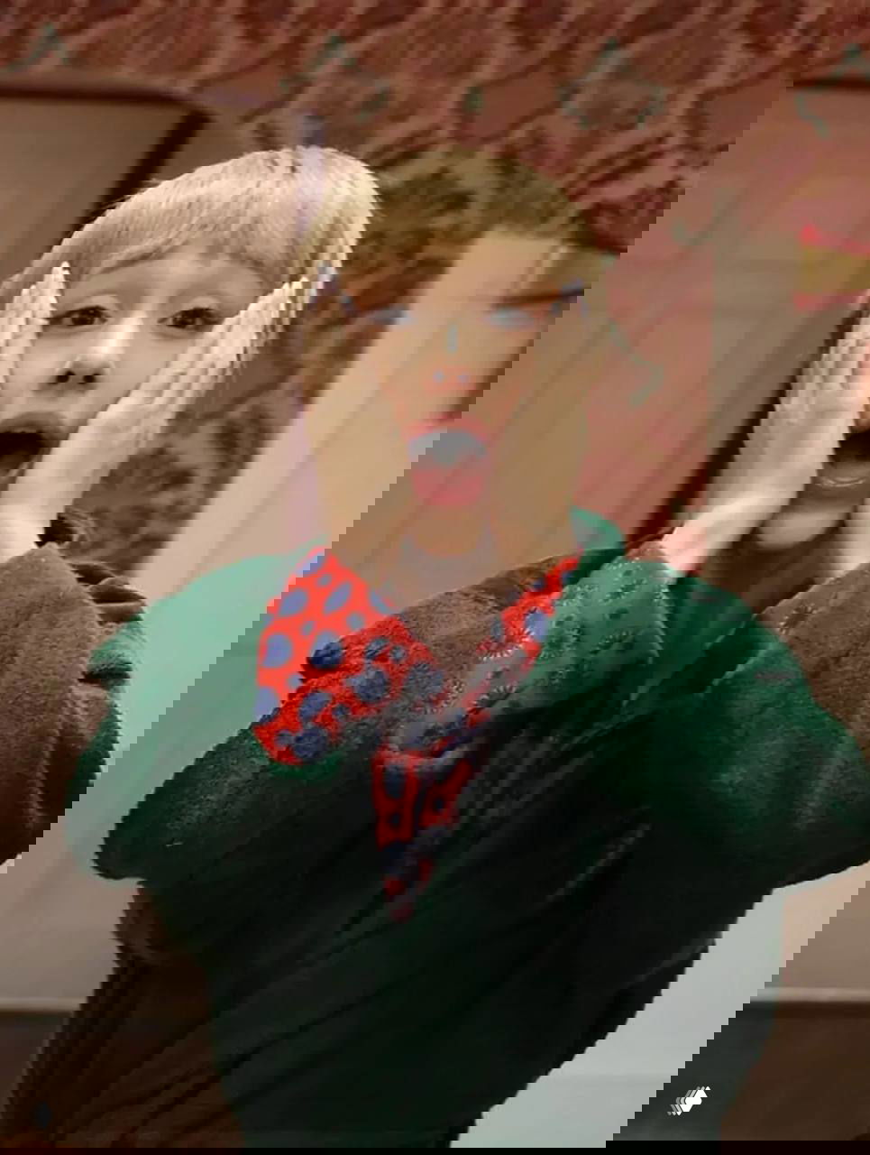 Крупный план женщины в зелёном халате, руки прижаты к щекам в классической позе удивления, на фоне узорные обои и дверь.