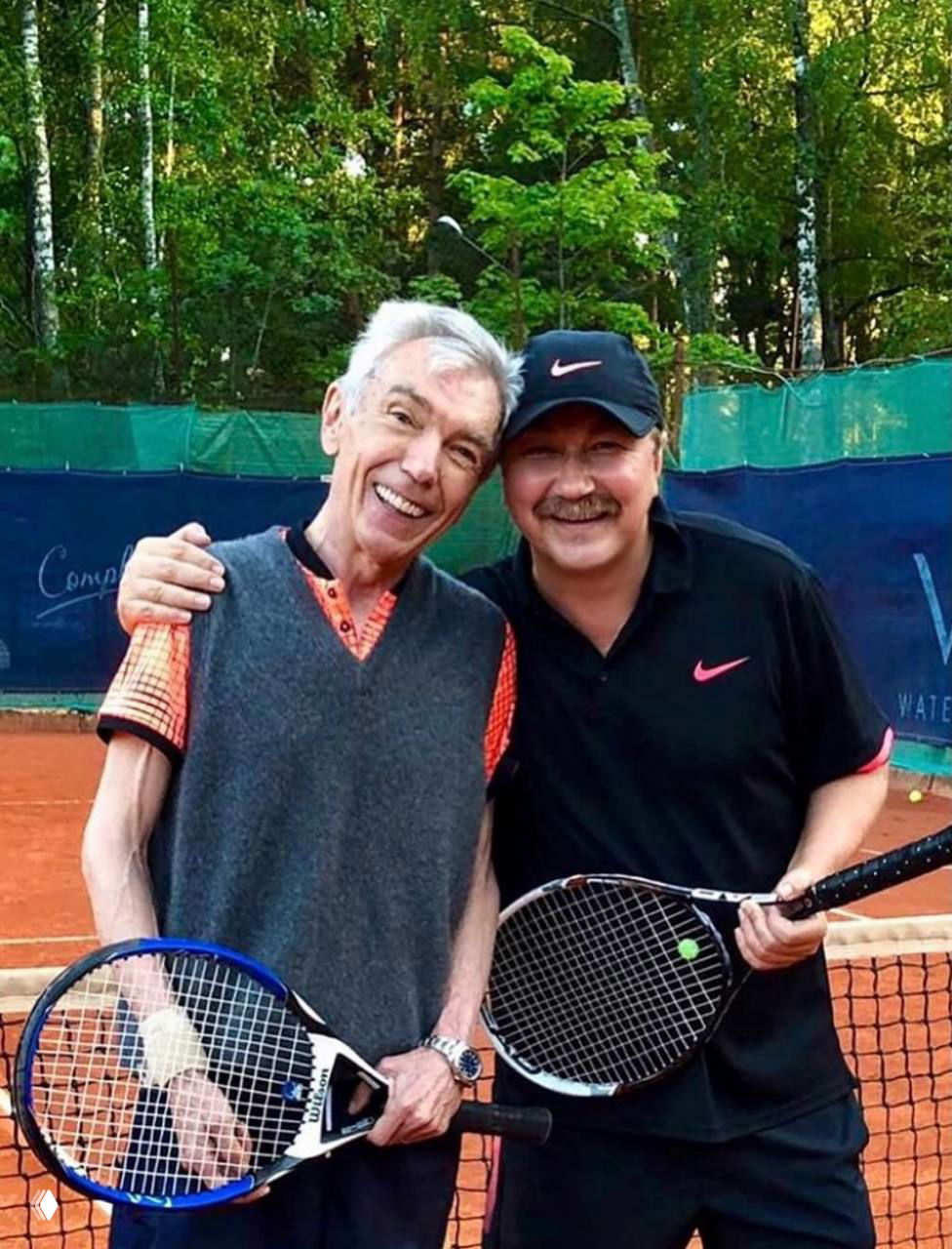 Двое улыбающихся мужчин стоят рядом на открытом теннисном корте, держат теннисные ракетки; зелёный лес и сетка корта видны на заднем плане.