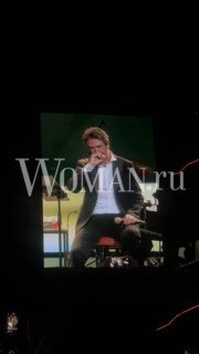 Исполнитель на сцене во время выступления, крупный экран с проекцией за ним и видимый логотип Woman.ru; темный концертный зал и микрофон