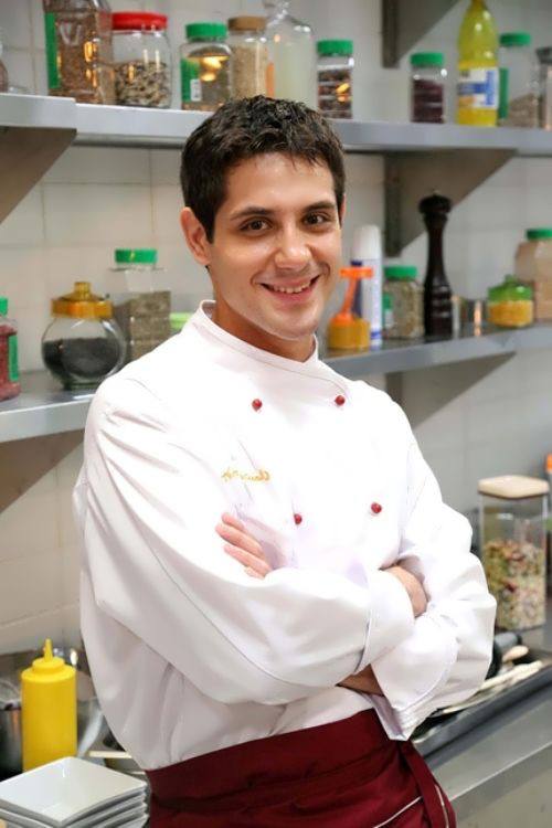 Кулинарный кадр: улыбающийся мужчина в белом колпаке и фартуке позирует в кухонной обстановке, профессиональная атмосфера ресторана или съёмочной кухни.