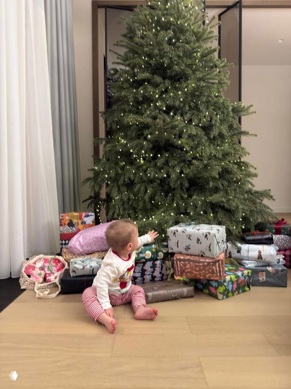 Малыш на полу у ёлки тянется к подаркам: много упакованных коробок у большой украшенной рождественской ели в домашнем интерьере.
