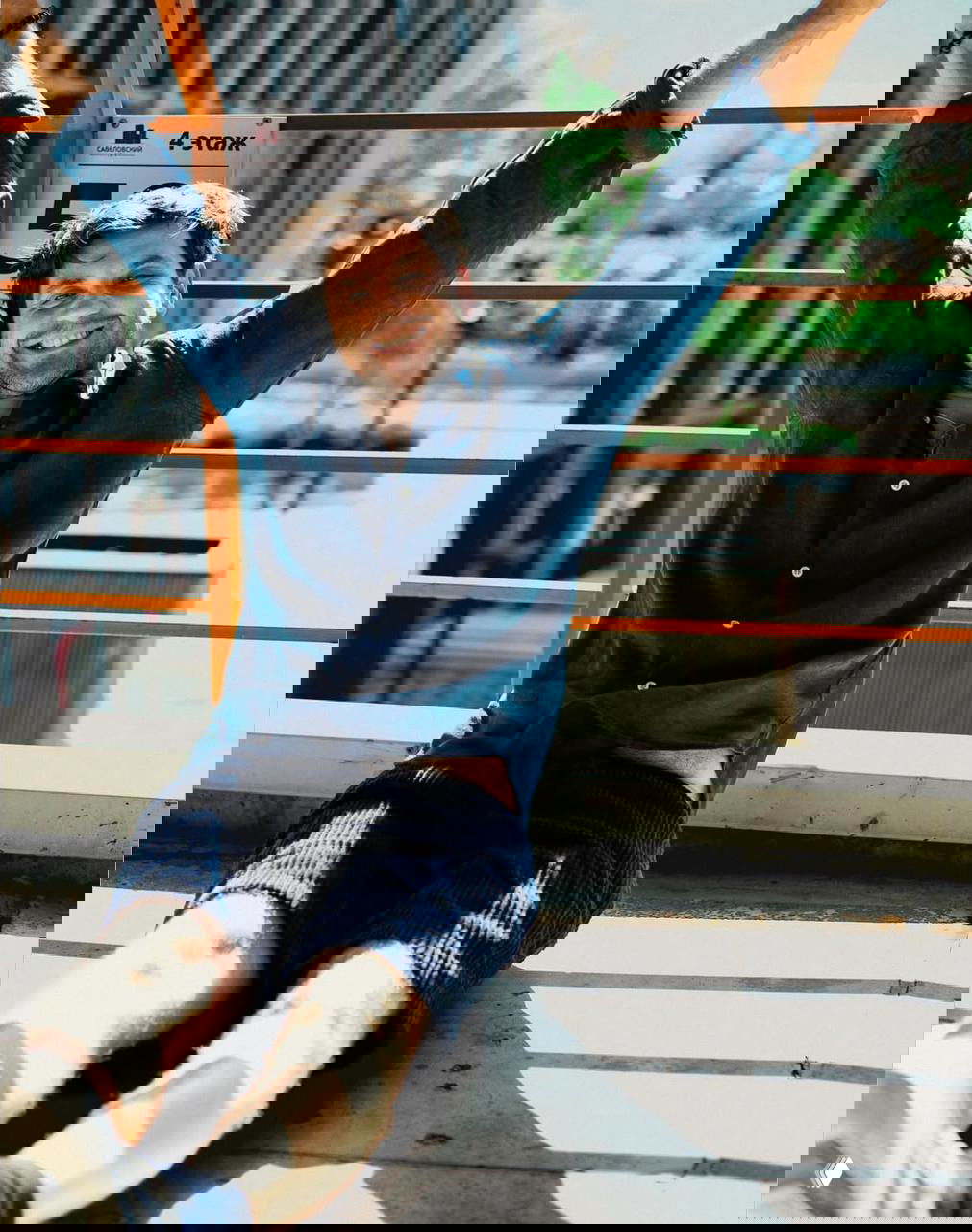 Мужчина в синей рубашке и шортах сидит на открытой площадке, тянет руки вверх и улыбается; летний непринуждённый портрет на балконе.