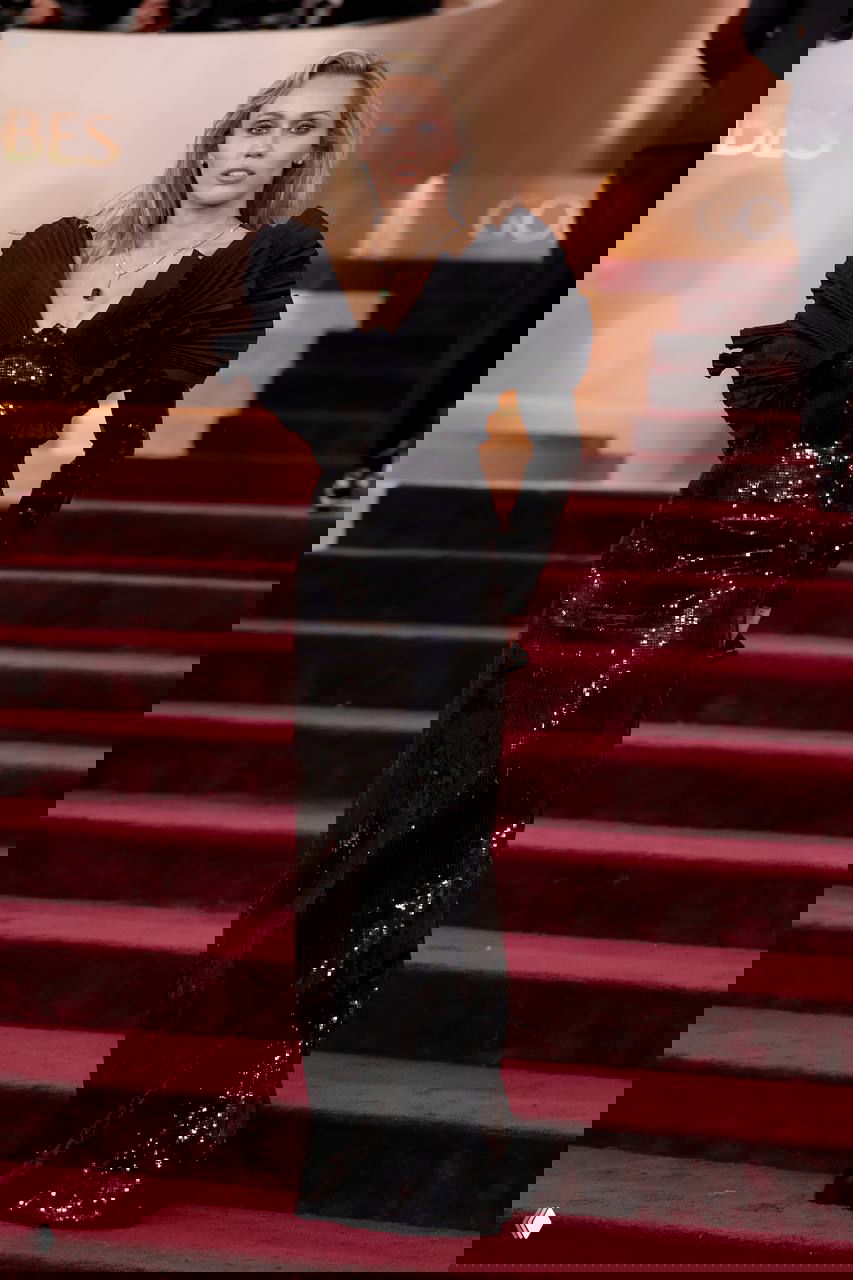 Женщина в блестящем чёрном платье с выразительными пышными плечами на ступенях красной дорожки; на заднем плане эмблема и операторы.