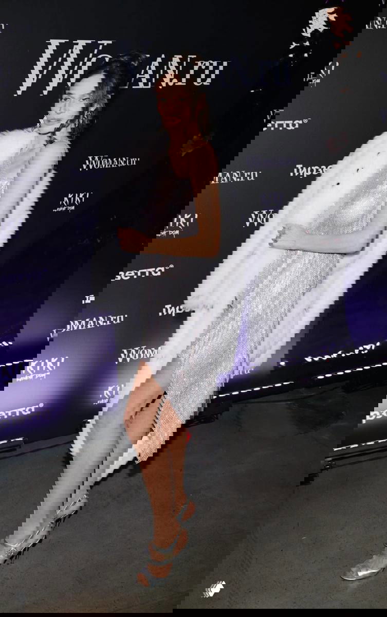 Женщина в блестящем серебристом платье с белой шубой позирует на фоне баннера Woman.ru во время вечернего мероприятия.