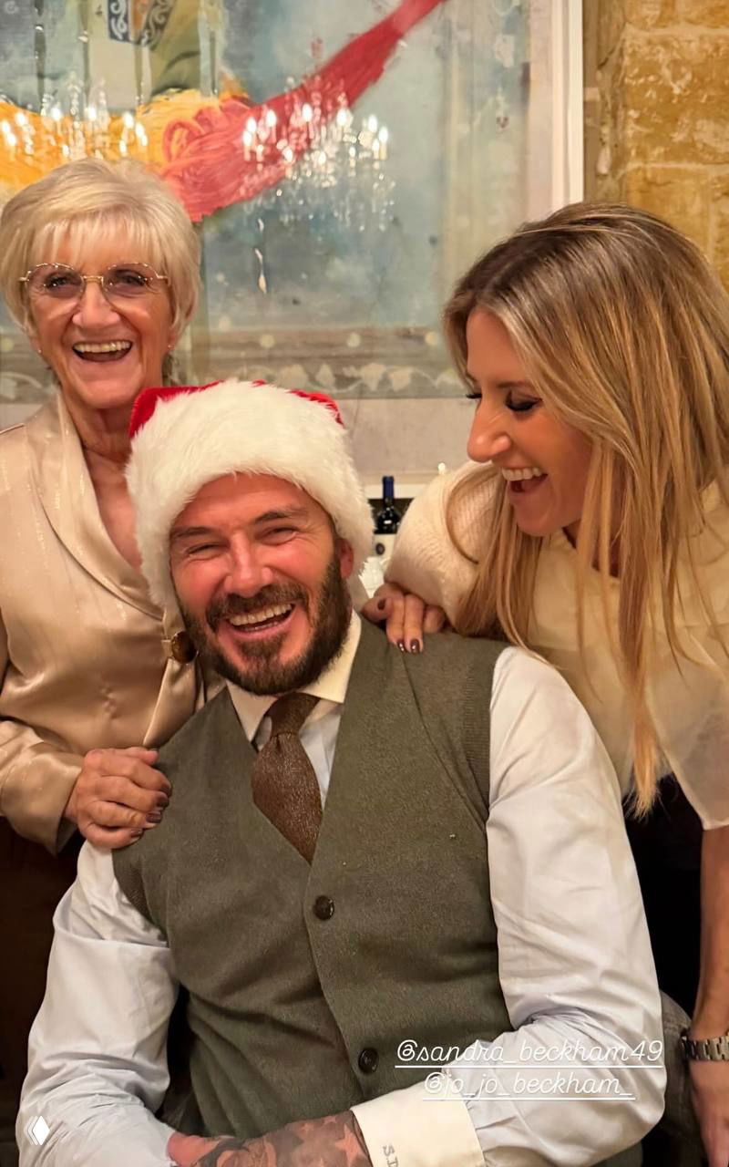 Несколько гостей улыбаются за праздничным столом: мужчина в шапке Санты и рядом женщины, праздничное настроение и смех.