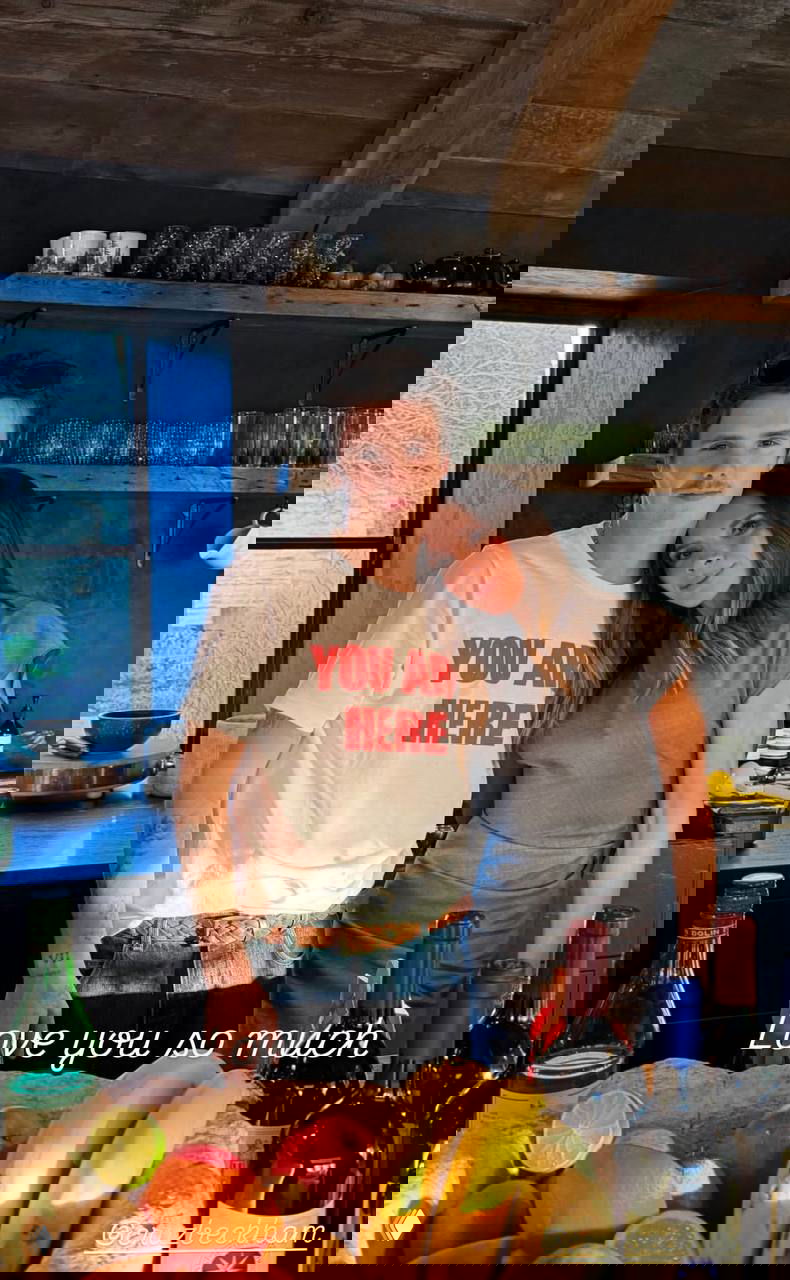 Двое позируют на кухне за столом с фруктами и бутылками вина; надпись на фото «Love you so much», праздничная домашняя сцена.