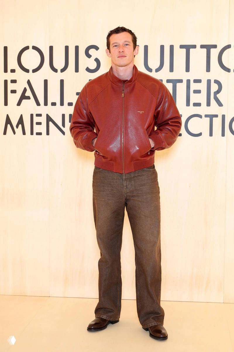 Мужчина в коричнево-красной кожаной куртке и прямых брюках на светлом фоне с логотипом бренда — минималистичный casual-образ для показа.