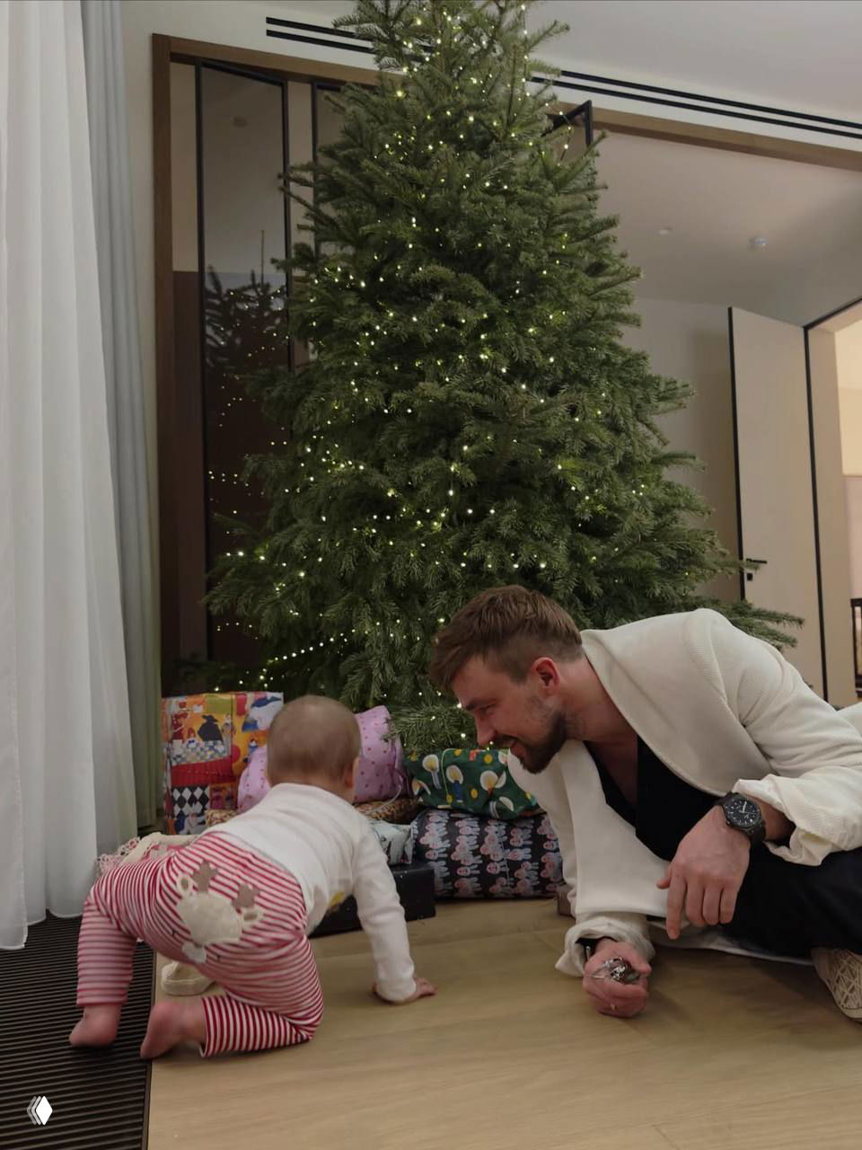 Александр играет с ребёнком у ёлки: мужчина на полу улыбается малышу, рядом множество красиво упакованных подарков и огоньки гирлянды.