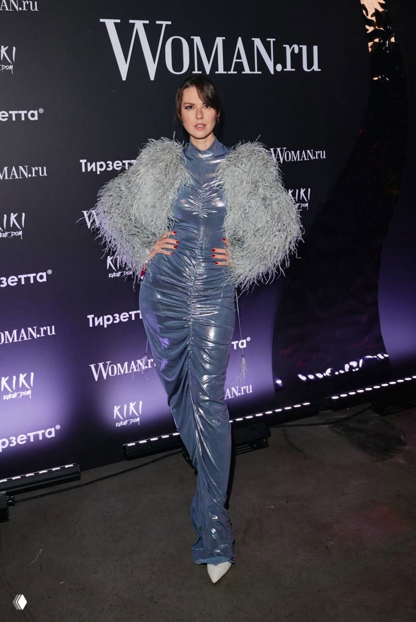 Женщина в эффектном длинном платье с пушистыми рукавами позирует у брендволла Woman.ru на вечерней вечеринке.