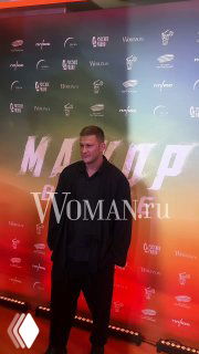 Мужчина в тёмной одежде позирует на красной дорожке у бренд-стены с логотипами и крупным названием фильма «Мажор» в вечерней подсветке.