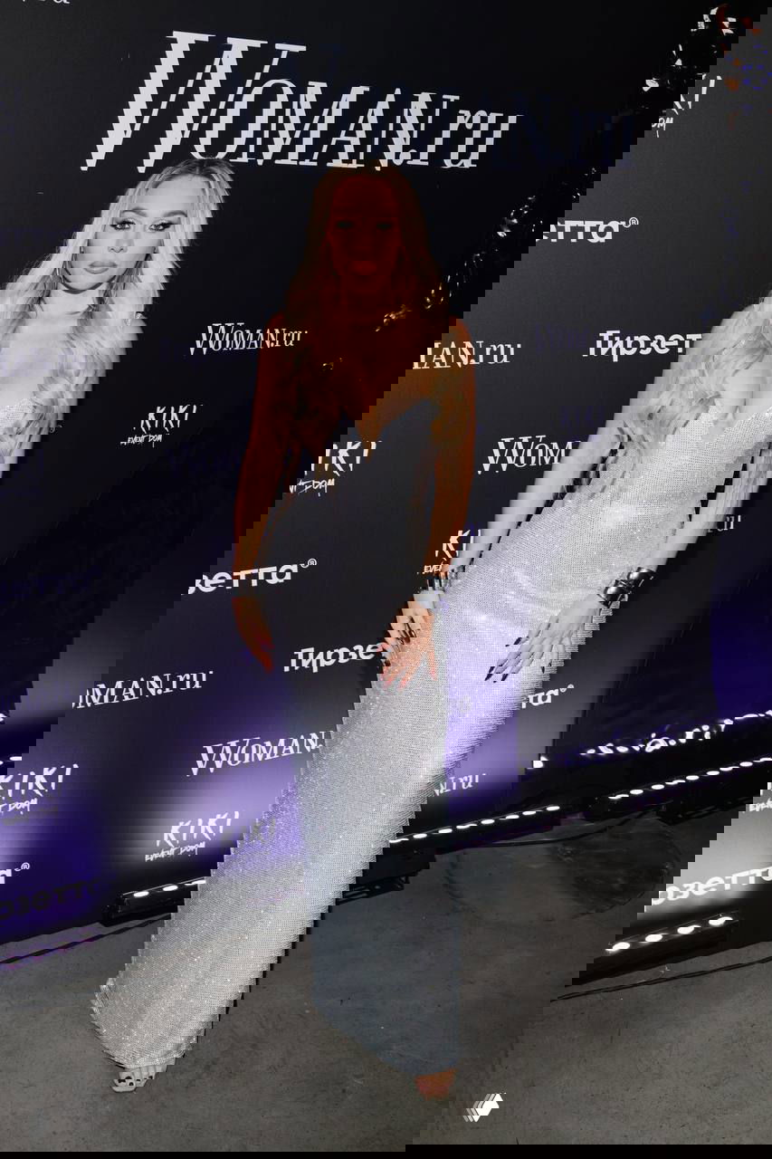 Женщина в длинном серебристом платье с декольте позирует на фоне логотипа Woman.ru в фотозоне мероприятия.