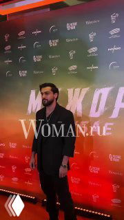 Мужчина в тёмной рубашке и пиджаке позирует на фоне баннера с брендингом фильма, сцена украшена цветной подсветкой.