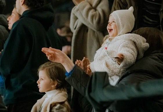 Несколько детей в тёплой одежде и шапках сидят на трибуне и с восторженным вниманием смотрят ледовое шоу — праздничная семейная сцена.