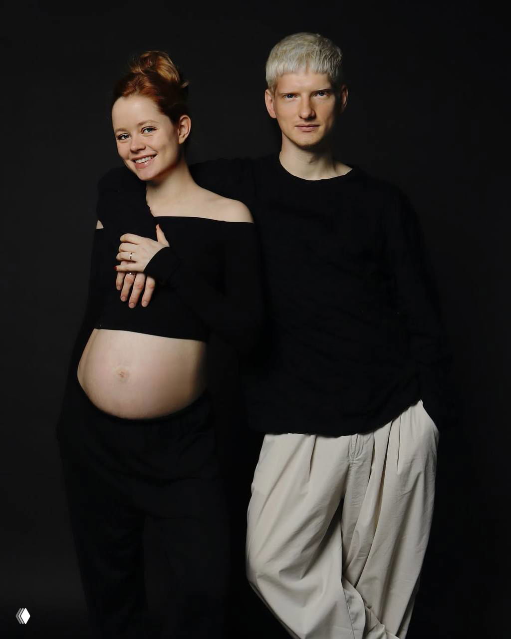 Мила Ершова улыбается рядом со Святославом Рогожаном на студийном тёмном фоне; актриса показывает живот — семейная фотосессия.