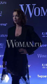 Гостья в чёрном жакете позирует на красной дорожке у баннера Woman.ru на вечеринке в честь дня рождения издания Woman.ru.