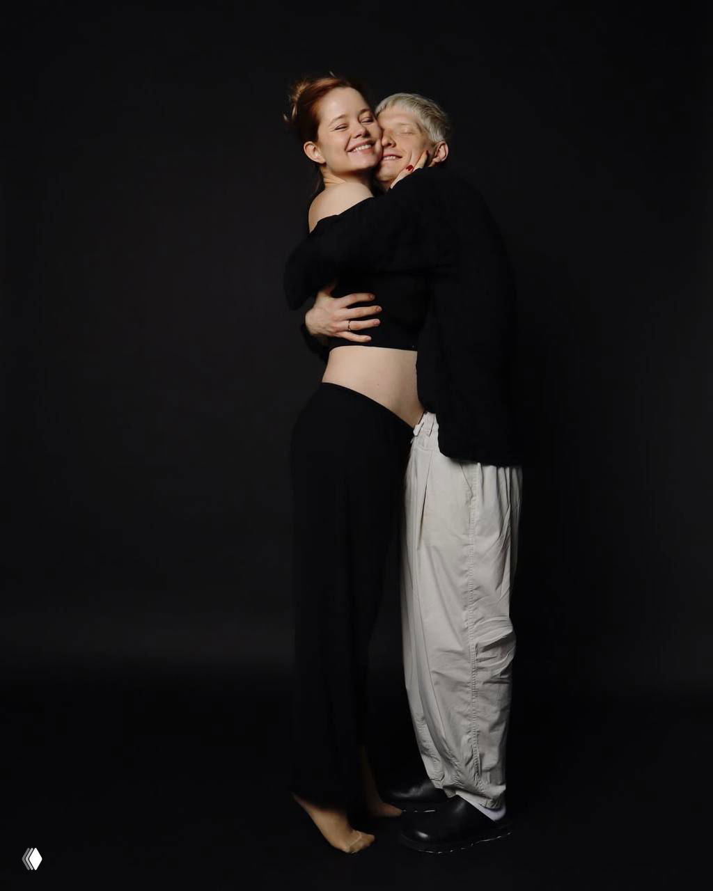 Мила Ершова и Святослав Рогожан крепко обнялись на чёрном фоне; снимок подчёркивает радость пары и ожидание ребёнка.
