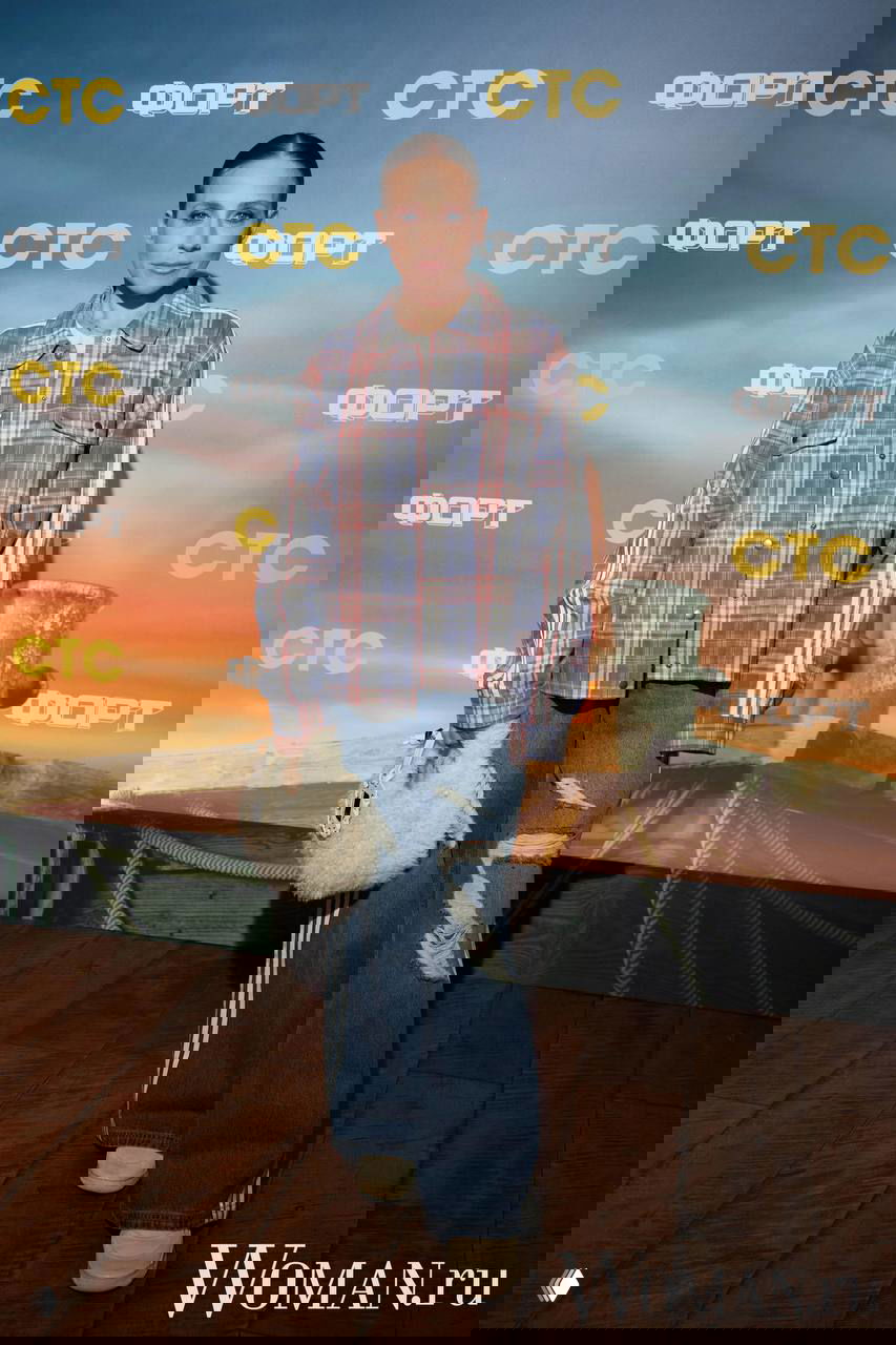 Женщина в свободной клетчатой рубашке и широких рваных джинсах позирует на фоне постера премьеры шоу, фон с логотипами СТС