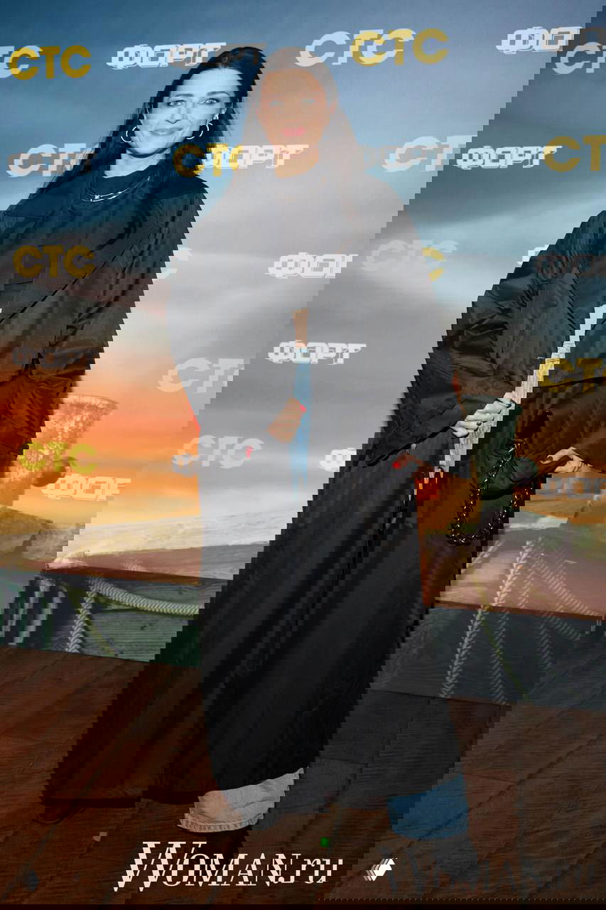 Женщина в длинном тёмном плаще и джинсах позирует на ковровой дорожке премьеры «Форт. Возвращение легенды», на фоне логотипы СТС и брендовый баннер