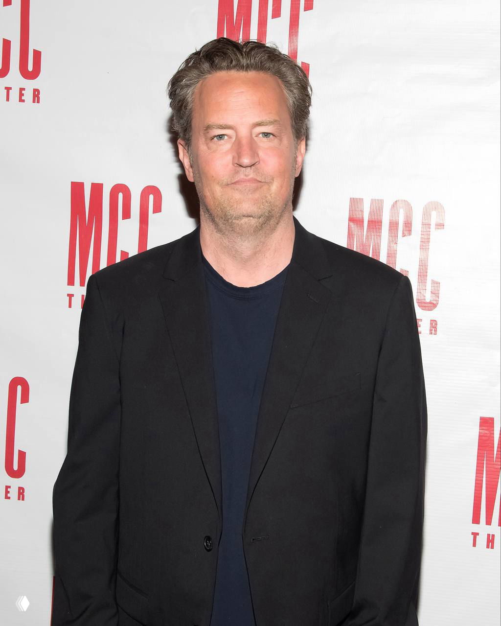 Мужчина средних лет в тёмном пиджаке и футболке на фоне белого баннера с крупными красными буквами, кадр с публичного события