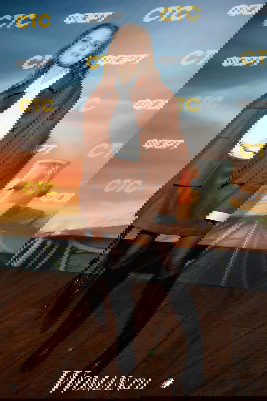 Женщина в коричневом костюме и высоких сапогах стоит на ковровой дорожке премьеры, позирует на фоне баннера с логотипами шоу и телеканала