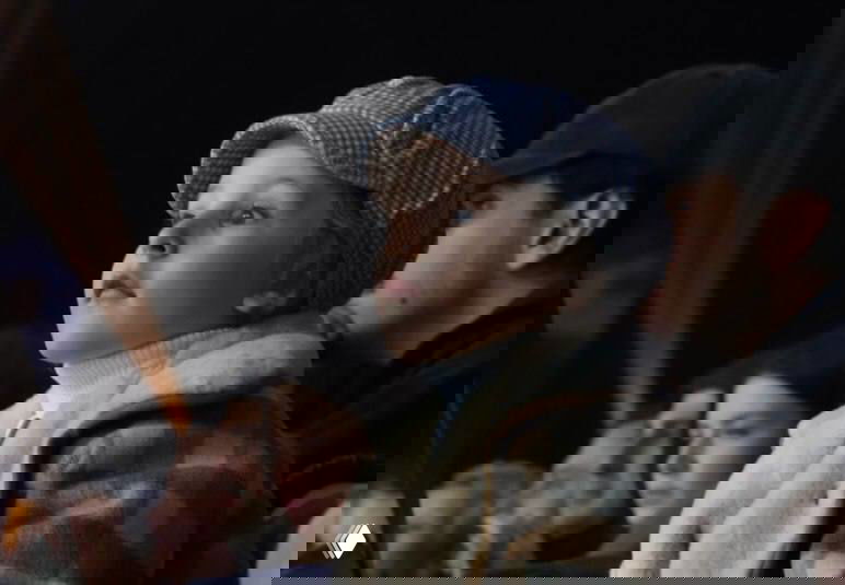 Ребёнок в кепке и тёплой одежде внимательно и серьёзно смотрит на сцену, в глубине кадра видны другие зрители и вечерняя подсветка.