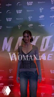 Молодая женщина в повседневном наряде выходит на красную дорожку перед фотоколлом с логотипами и названием картины на заднем плане.