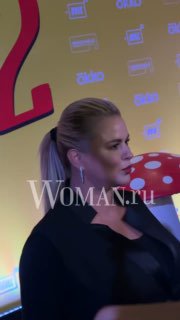 Анна Семенович в профиль на мероприятии: светлая причёска хвостом, тёмная одежда, на заднем плане желтая декорация с логотипом и видимым водяным знаком Woman.ru
