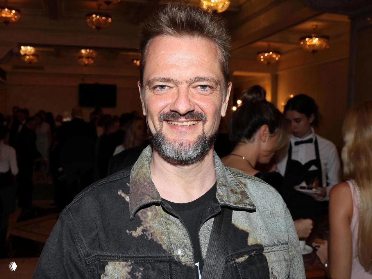 Улыбающийся телеведущий в джинсовой куртке на светском приёме: крупный план лица, в зале видны люстры и размытая толпа гостей на фоне.