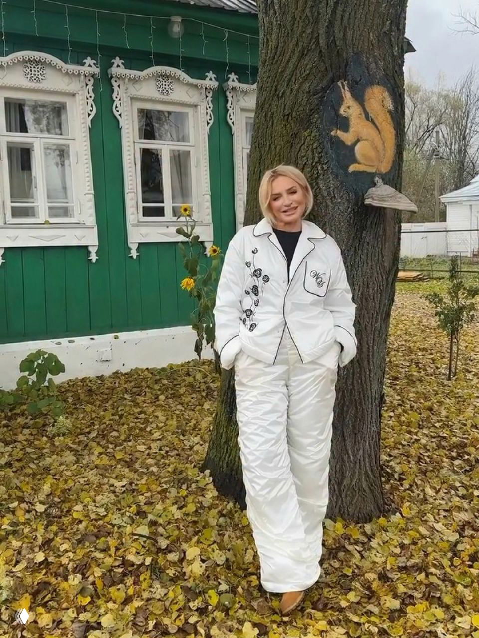 Женщина в белом стёганом костюме позирует у дерева с изображением белки и деревянного дома, осенний ковёр из листьев под ногами.