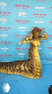 Хайди Клум позирует в образе Медузы Горгоны на синей фотозоне: вид в полный рост с акцентом на хвост и текстуру костюма.