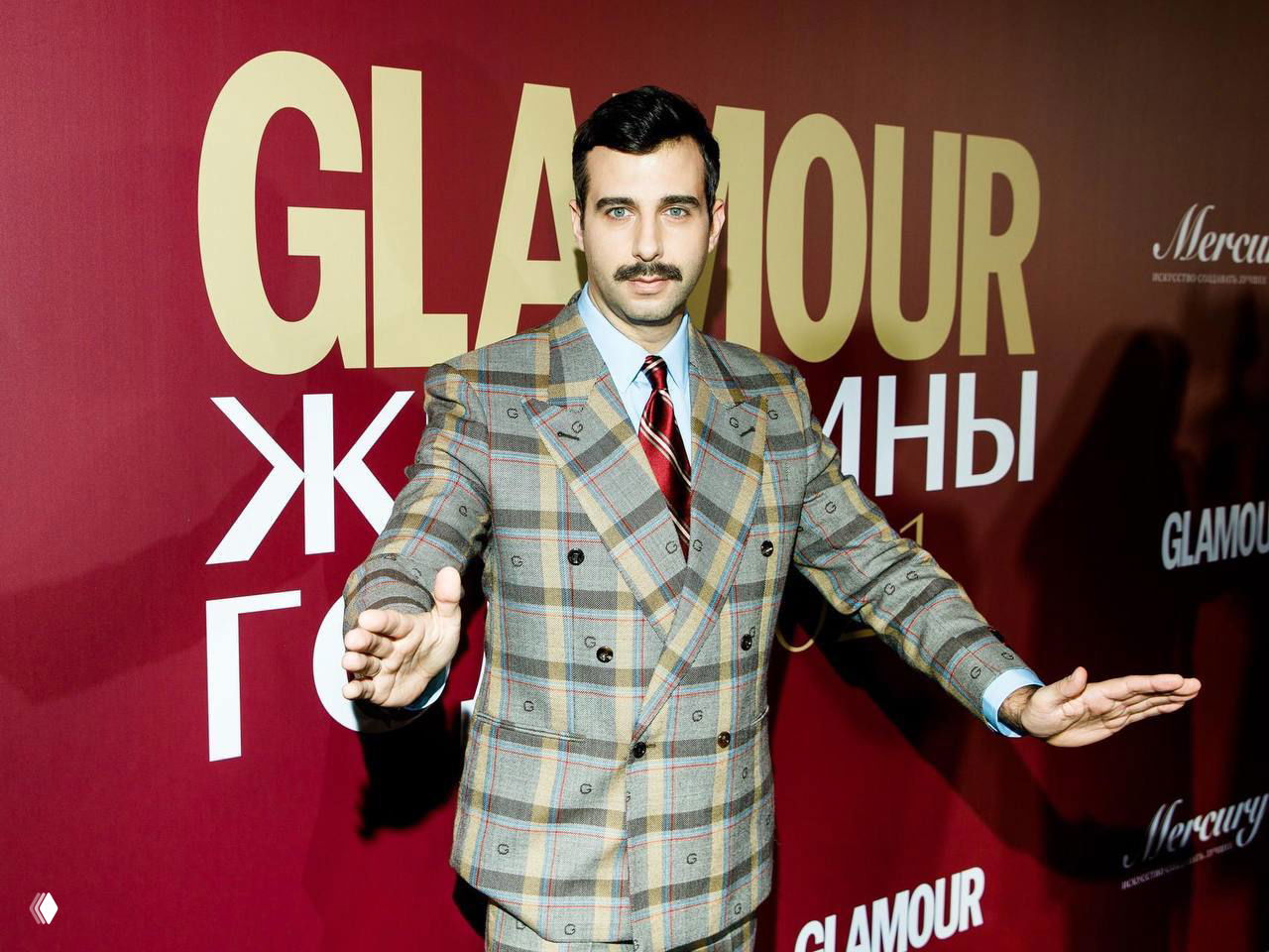 Телеведущий в клетчатом двубортном костюме и галстуке позирует на красной дорожке у баннера Glamour, делает жесты руками в сторону камеры.