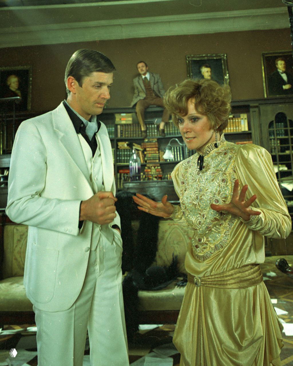 Сценический кадр: женщина в золотистом платье ведёт диалог с мужчиной в белом костюме на фоне книжного интерьера, кадр из фильма.