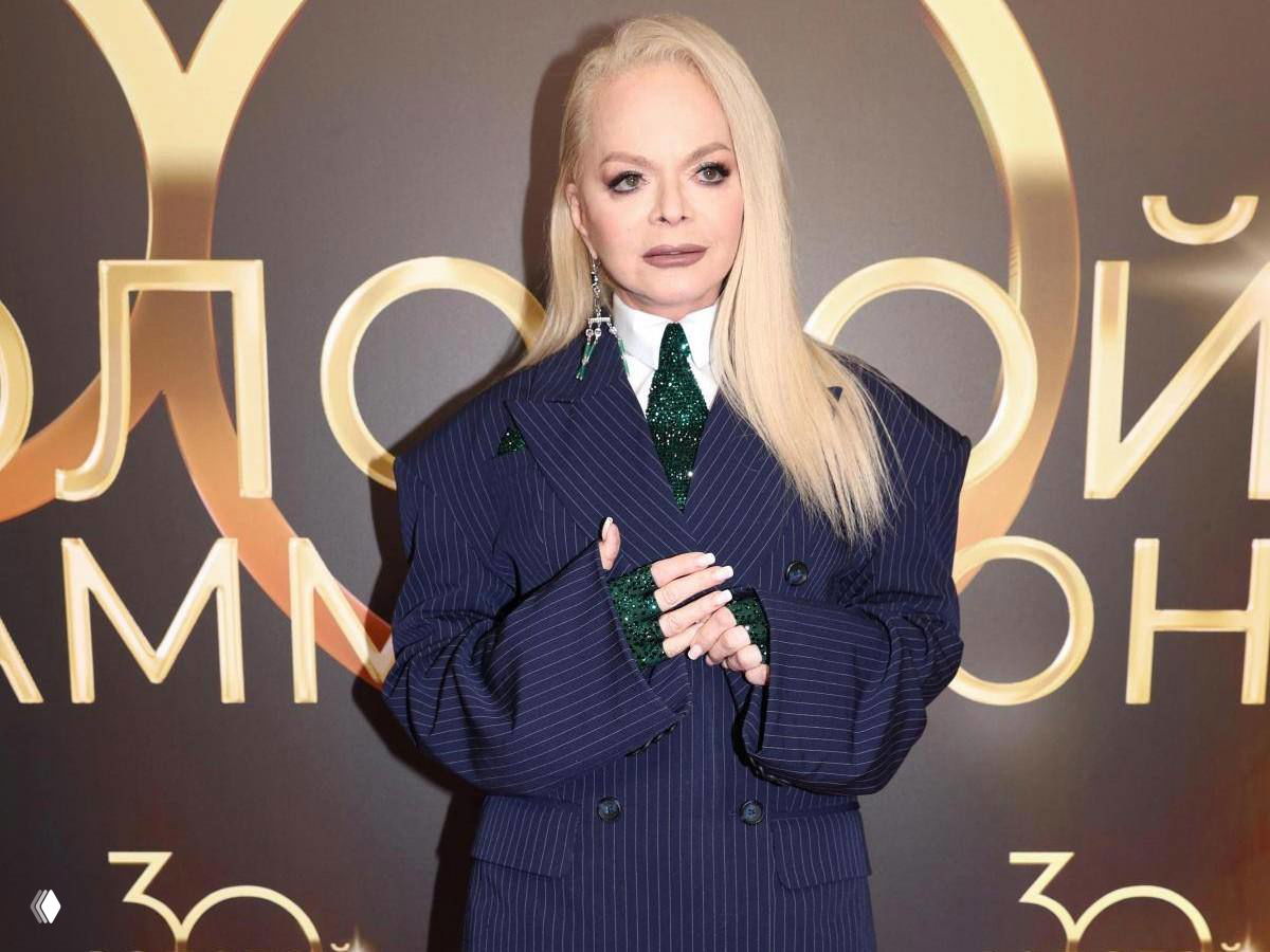 Женщина со светлыми волосами в тёмно-синем костюме позирует у брендированной фотозоны на светском мероприятии.