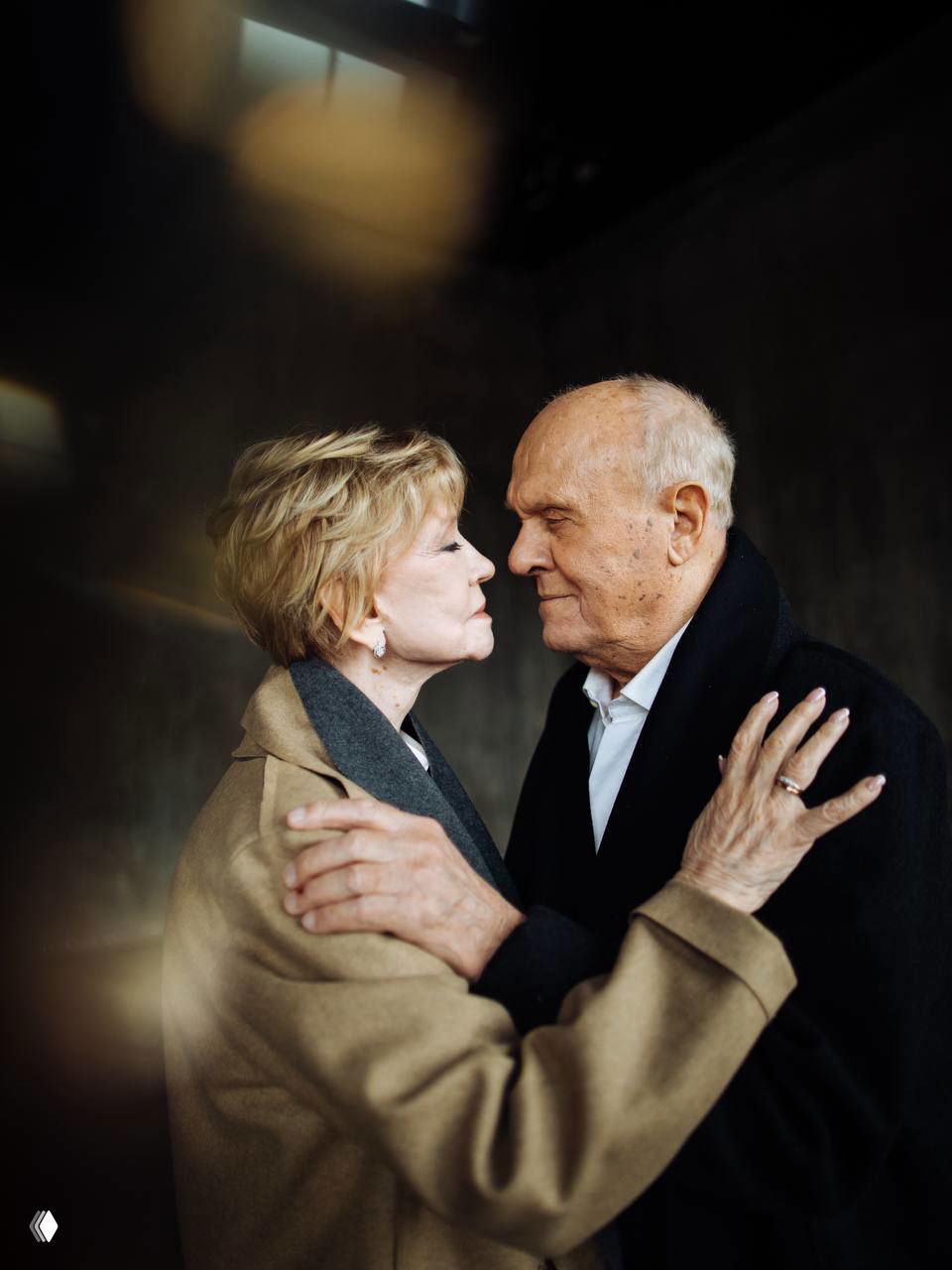 Боковой план пожилой пары, стоящей лицом друг к другу в приглушённом свете; кадр выражает интимность, уважение и тихую скорбь.