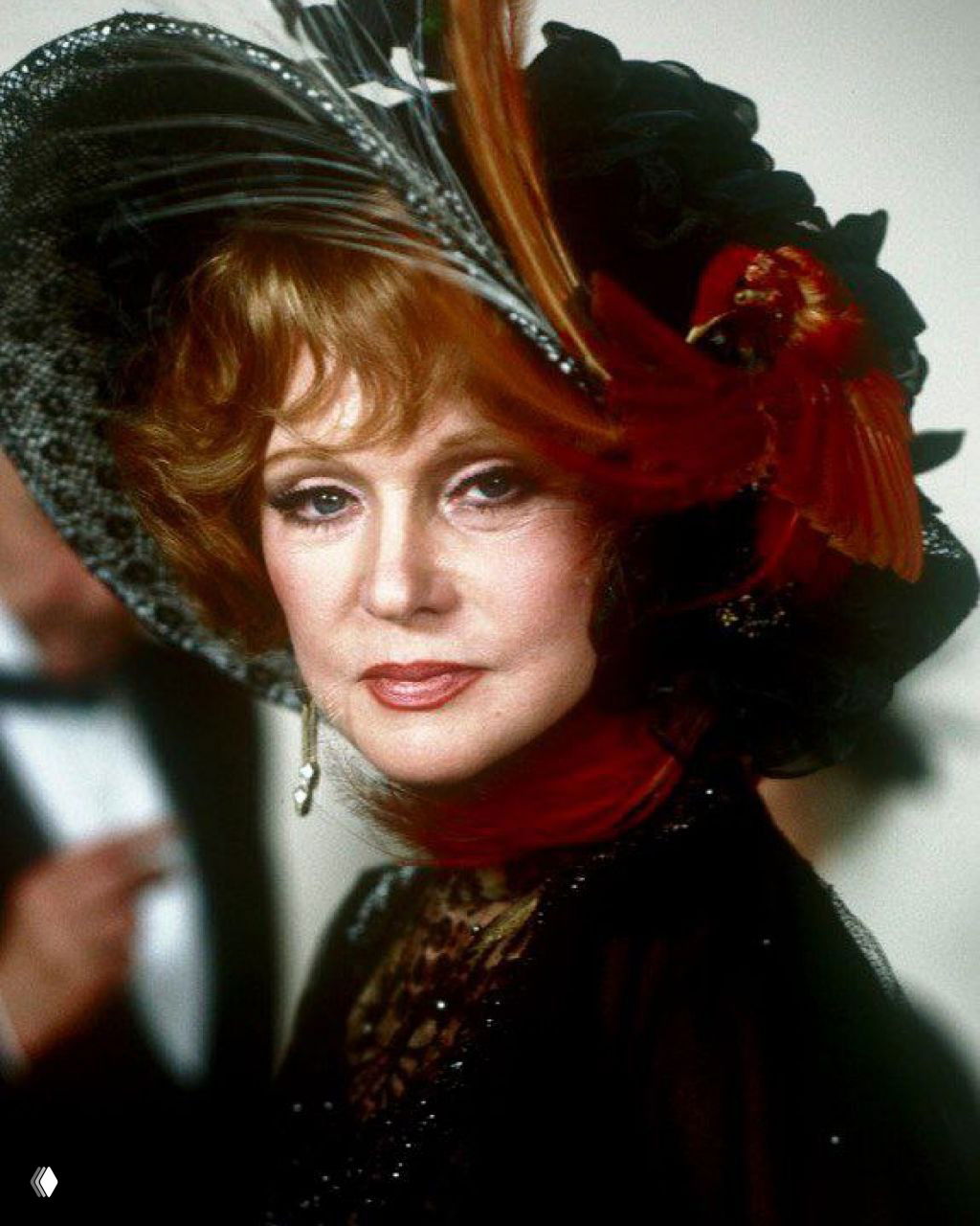 Крупный портрет в эффектной шляпе с перьями и ярким макияжем; театральный образ и декоративный реквизит, студийная фотография.