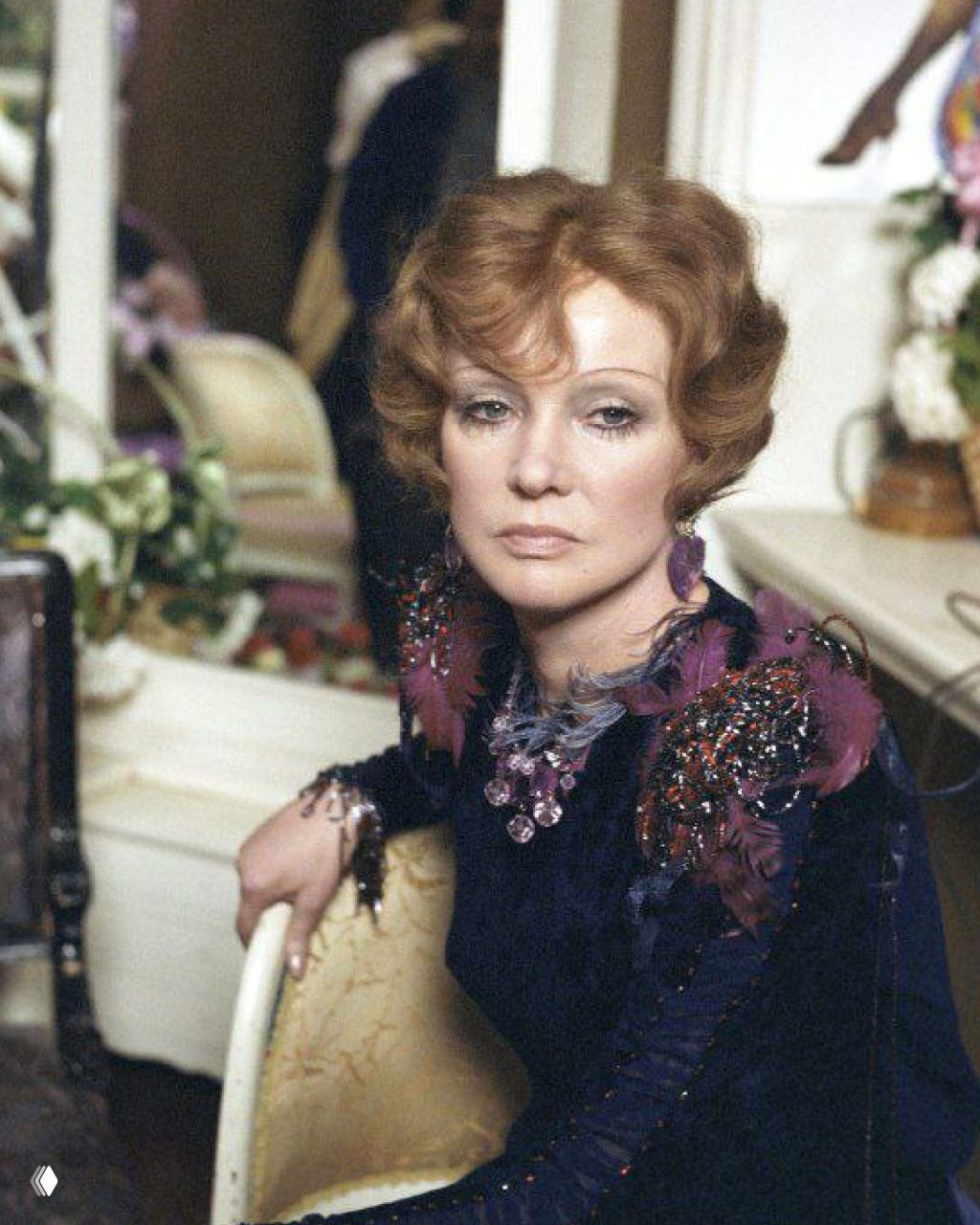 Кадр с задумчивым портретом женщины в роскошном платье и крупными украшениями, интерьер со шторами и классической мебелью на заднем плане.