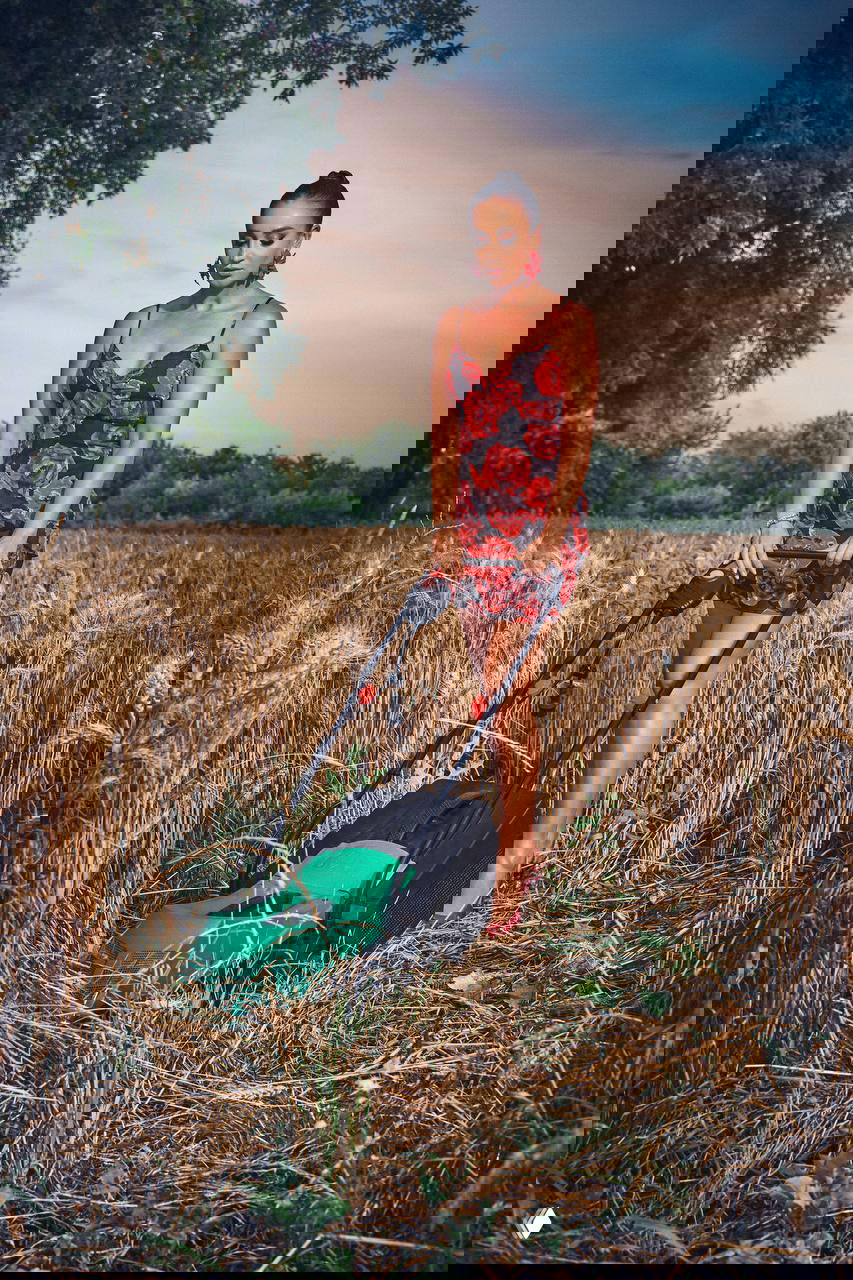 Ольга Бузова в ярком летнем платье и туфлях толкает газонокосилку по высохшему пшеничному полю — контраст сельского пейзажа и гламура.