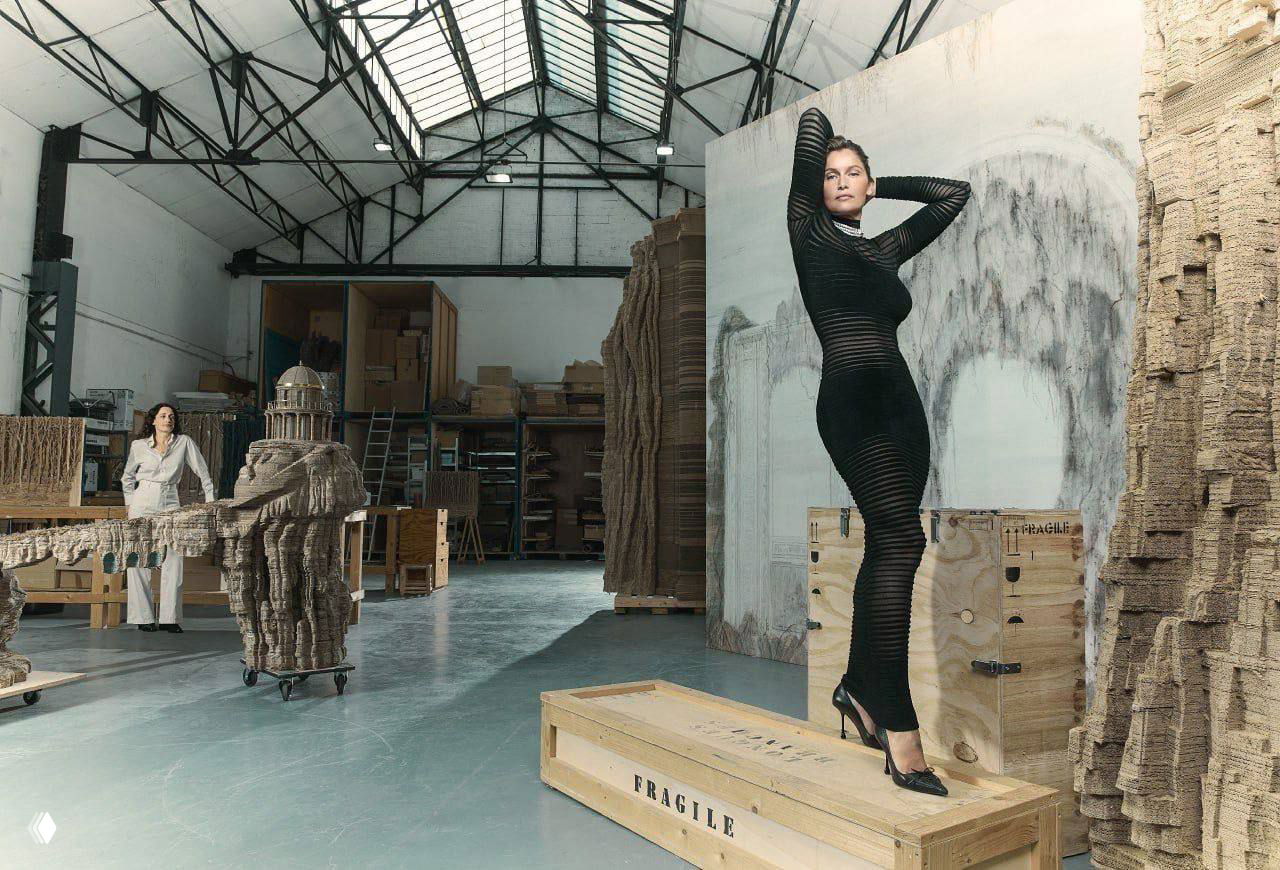Широкий план студии: модель в чёрном облегающем платье стоит на деревянном ящике среди арт-инсталляций и рабочих конструкций.