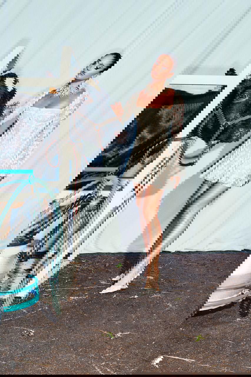 Ольга Бузова в коротком белом платье стоит рядом с коровой и доильным аппаратом в фермерском хлеве — фотосъёмка в сельской обстановке.