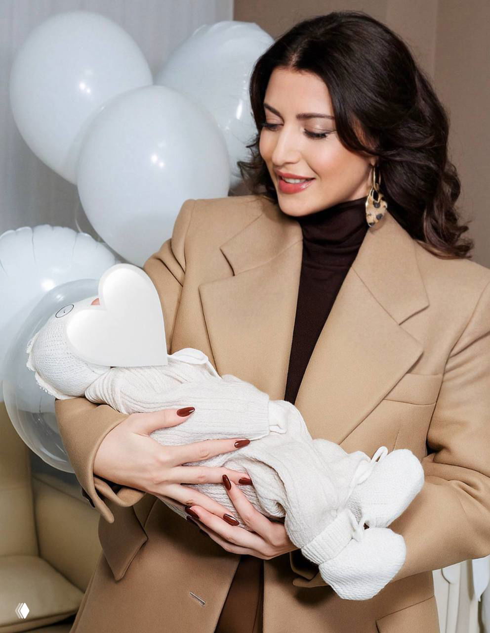 Жасмин в бежевом пальто держит на руках внука в тёплой одежде; лицо ребёнка закрыто стикером‑сердцем, на заднем плане белые шарики.