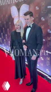 Дмитрий Дюжев в смокинге и его супруга в чёрном платье позируют вместе на красной дорожке премии «Бриллиантовая бабочка», фон с логотипами.