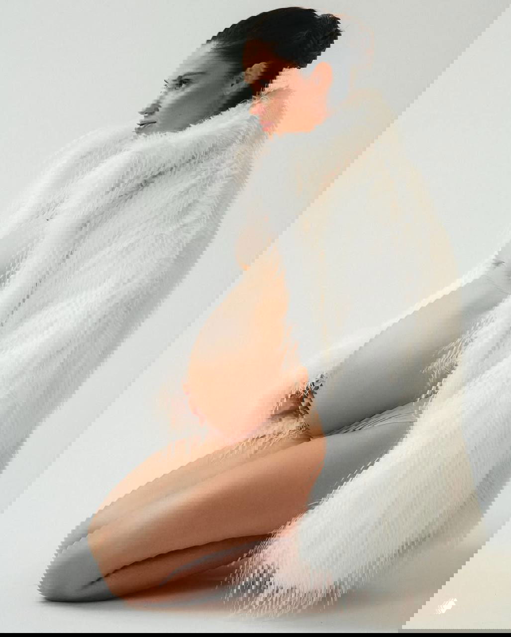 Беременная женщина в студийной съёмке: на корточках в бежевом белье и белой пушистой шубе на светлом фоне.