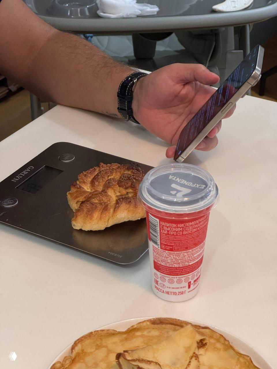 Рука держит смартфон над столом с круассаном на весах и стаканом йогурта — бытовой кадр с утренним перекусом и техникой рядом.