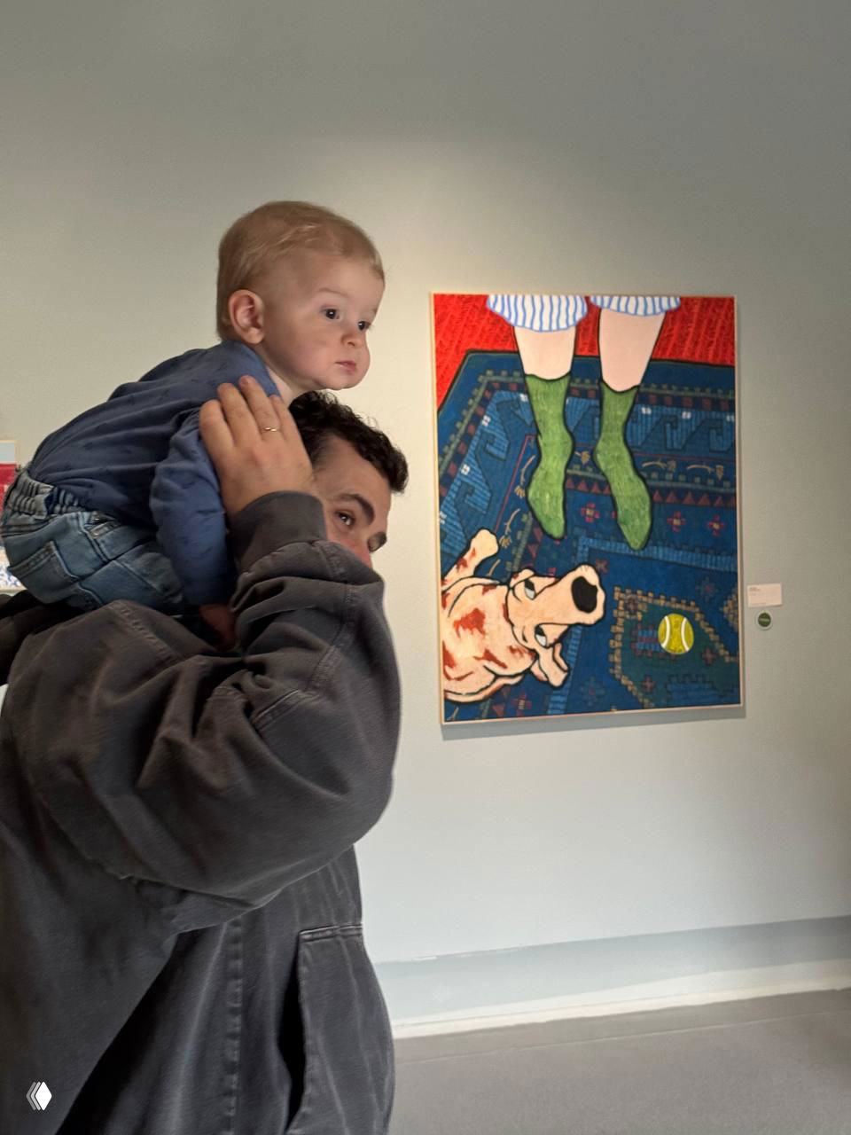 Мужчина несёт ребёнка на плечах в галерее рядом с яркой картиной и наблюдает экспозицию — семейный выход в музей или выставку.