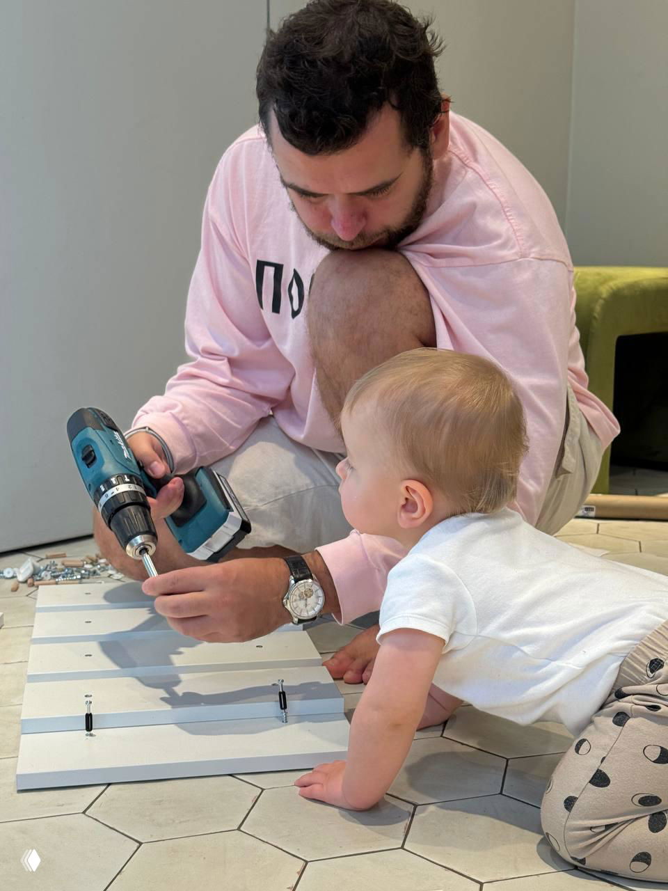 Отец использует шуруповёрт, а младенец ползает рядом и наблюдает — совместная сборка мебели или ремонт в доме с участием ребёнка.
