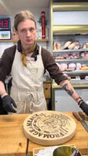 Продавец стоит за прилавком с большой деревянной доской и печатью, на заднем фоне полки и холодильники магазина.