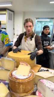 Молодой сотрудник режет большую головку сыра на деревянной разделочной доске, виден крупный план процесса и перчатки.