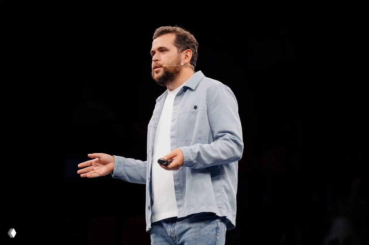 Спикер на сцене с наголовным микрофоном и пультом в руке, в светлой куртке и джинсах на тёмном фоне — репрезентативная фотография выступления.